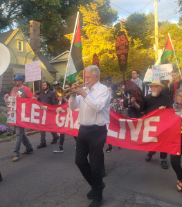 Man blowing trumpet in front of a line of marchers carrying a black banner with white writing saying 'Let Gaza Live'