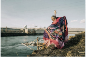 A photo of Lana Jack from behind, her arms spread out, a butterfly clip in her hair and wearing a black mult-color print, facing an industrial site on the river 